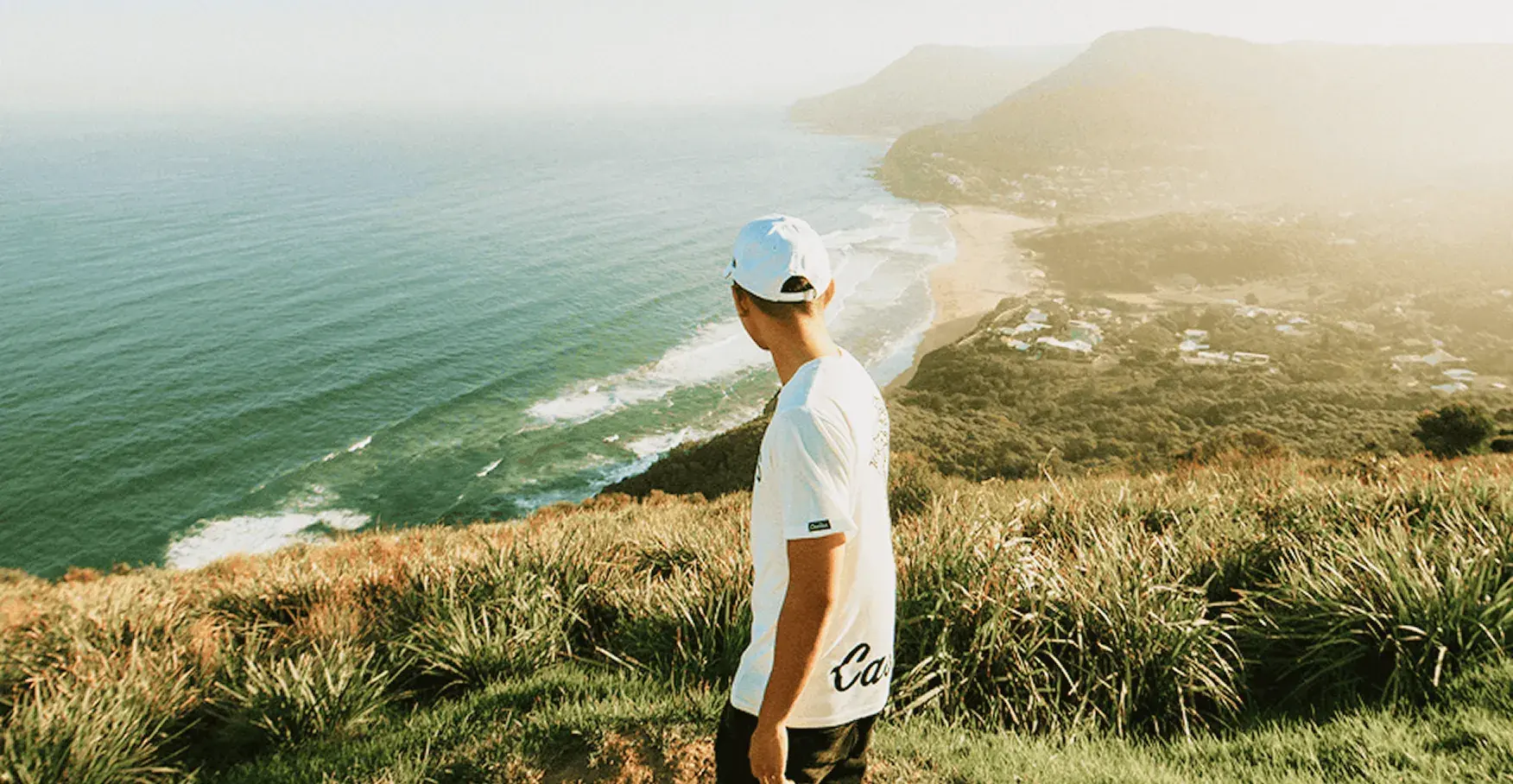 高台から海岸線を見下ろす、白いTシャツを着た男性の後ろ姿