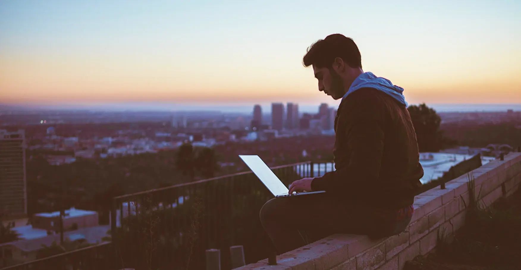 夕暮れの街を見渡す屋上の縁に座り、ノートパソコンを操作する男性