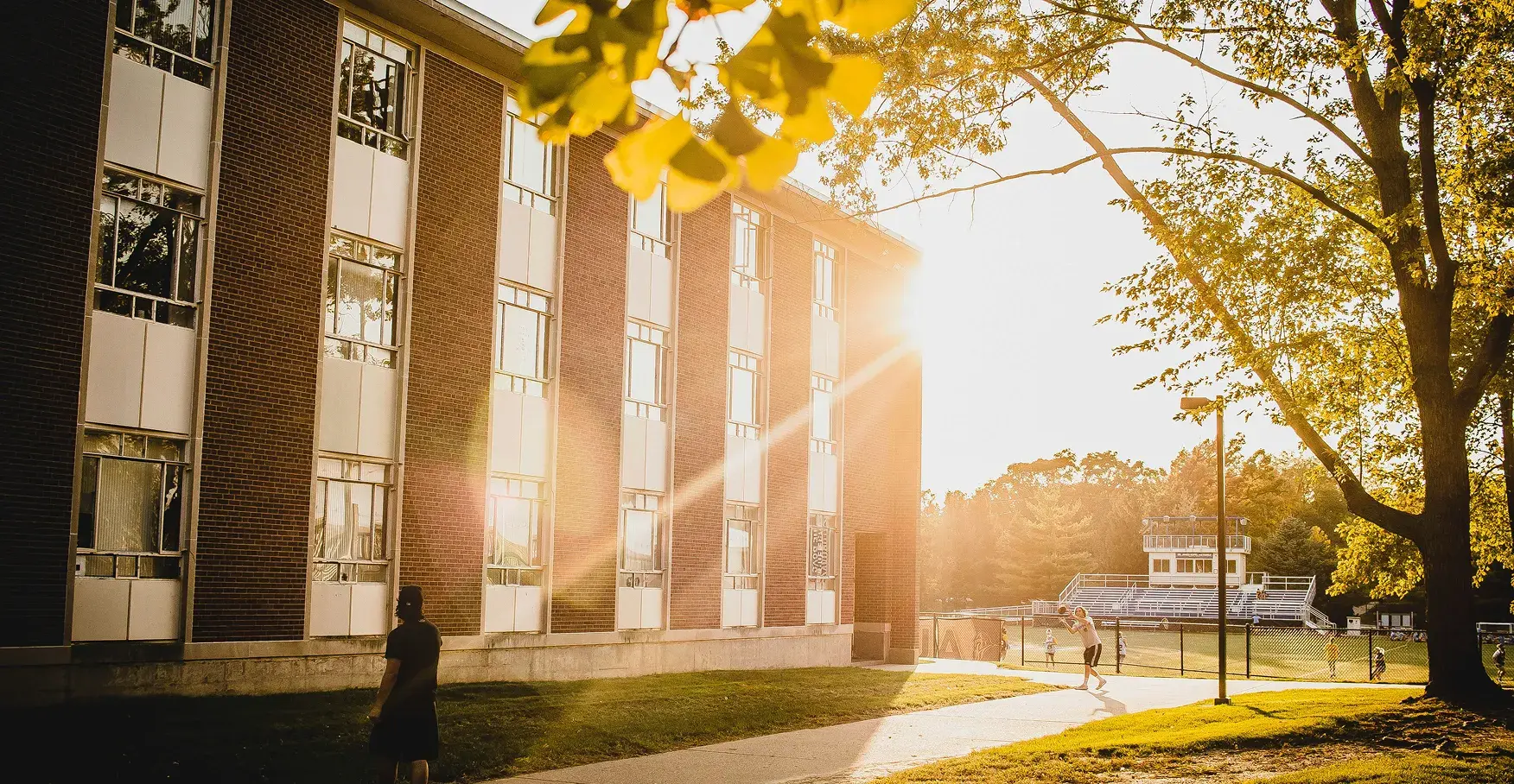夕日が差し込む大学のキャンパスで、レンガ造りの校舎の横を歩く学生たちと奥に見えるグラウンド
