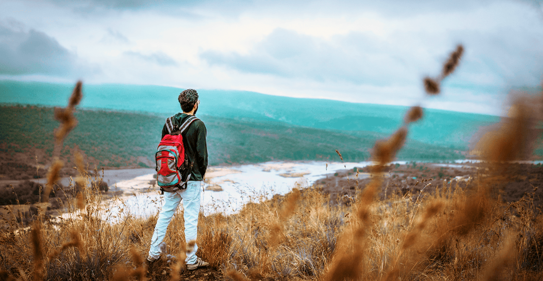 草原の縁でバックパックの青年が遠くの湖と山並みを見つめるイメージ