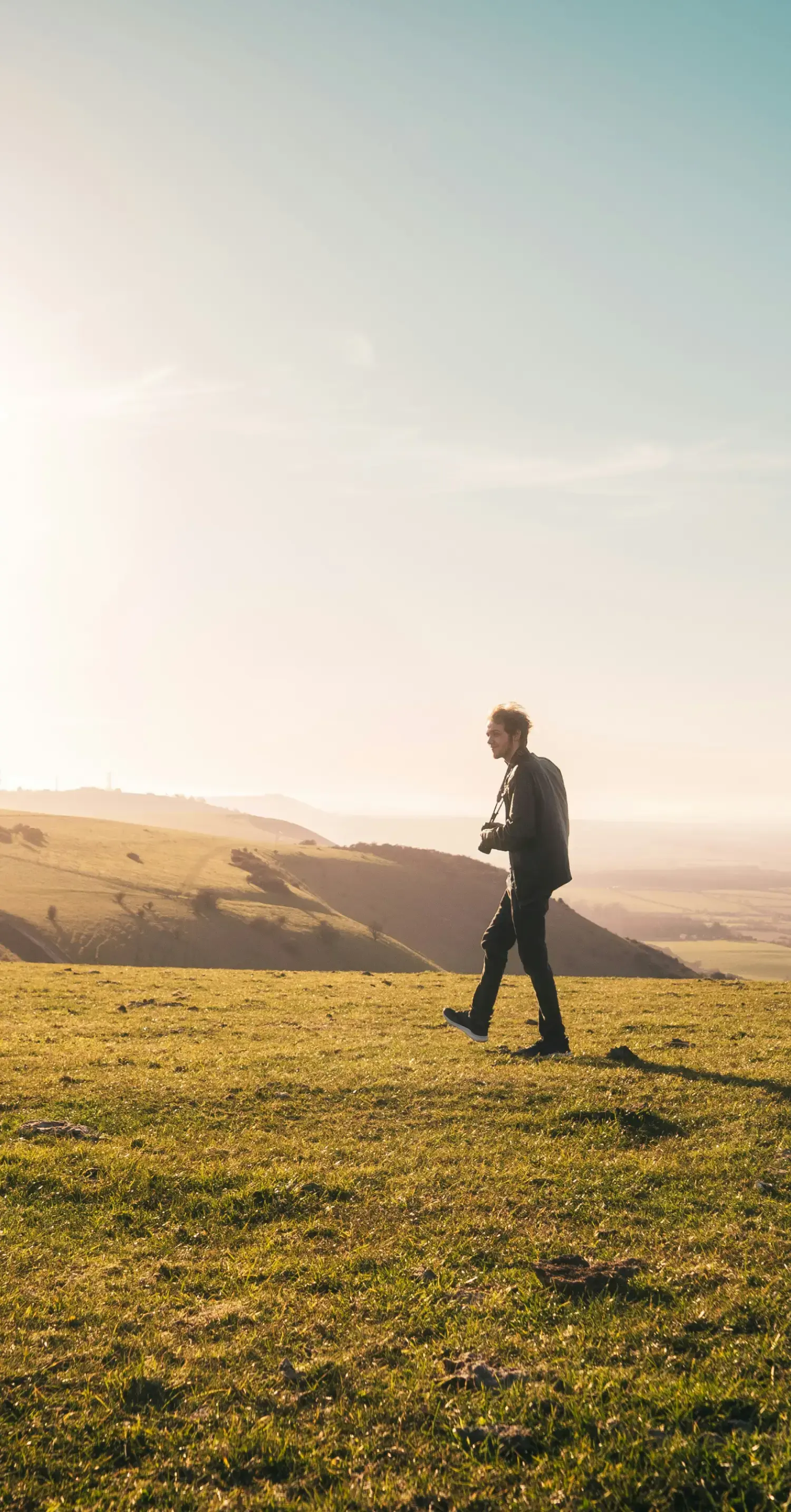 広大な丘陵地を歩く、カメラを下げた男性の横顔_メインビジュアル_sp