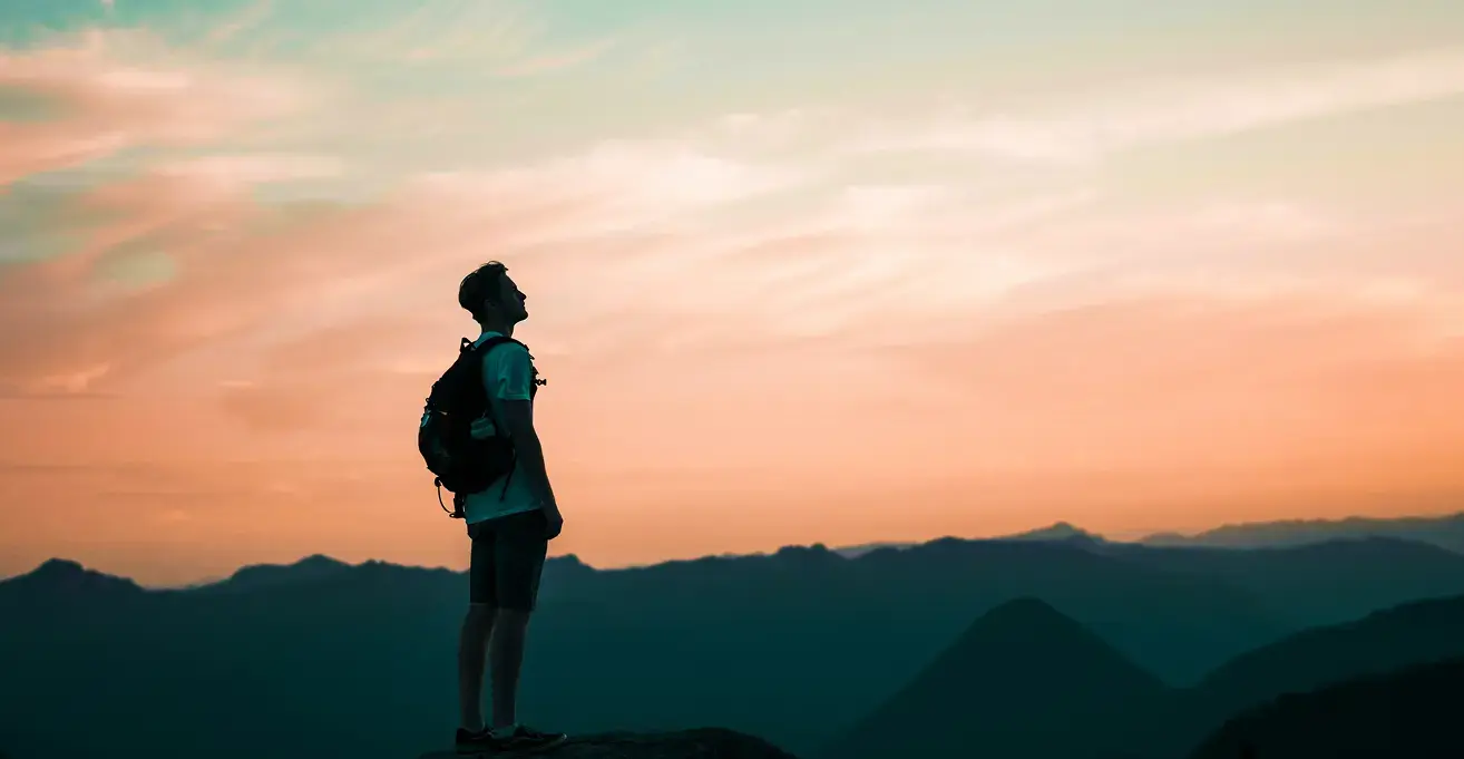 グラデーションに染まる美しい夕空の下、バックパックを背負い山頂から遠くを見つめるハイカーのシルエット